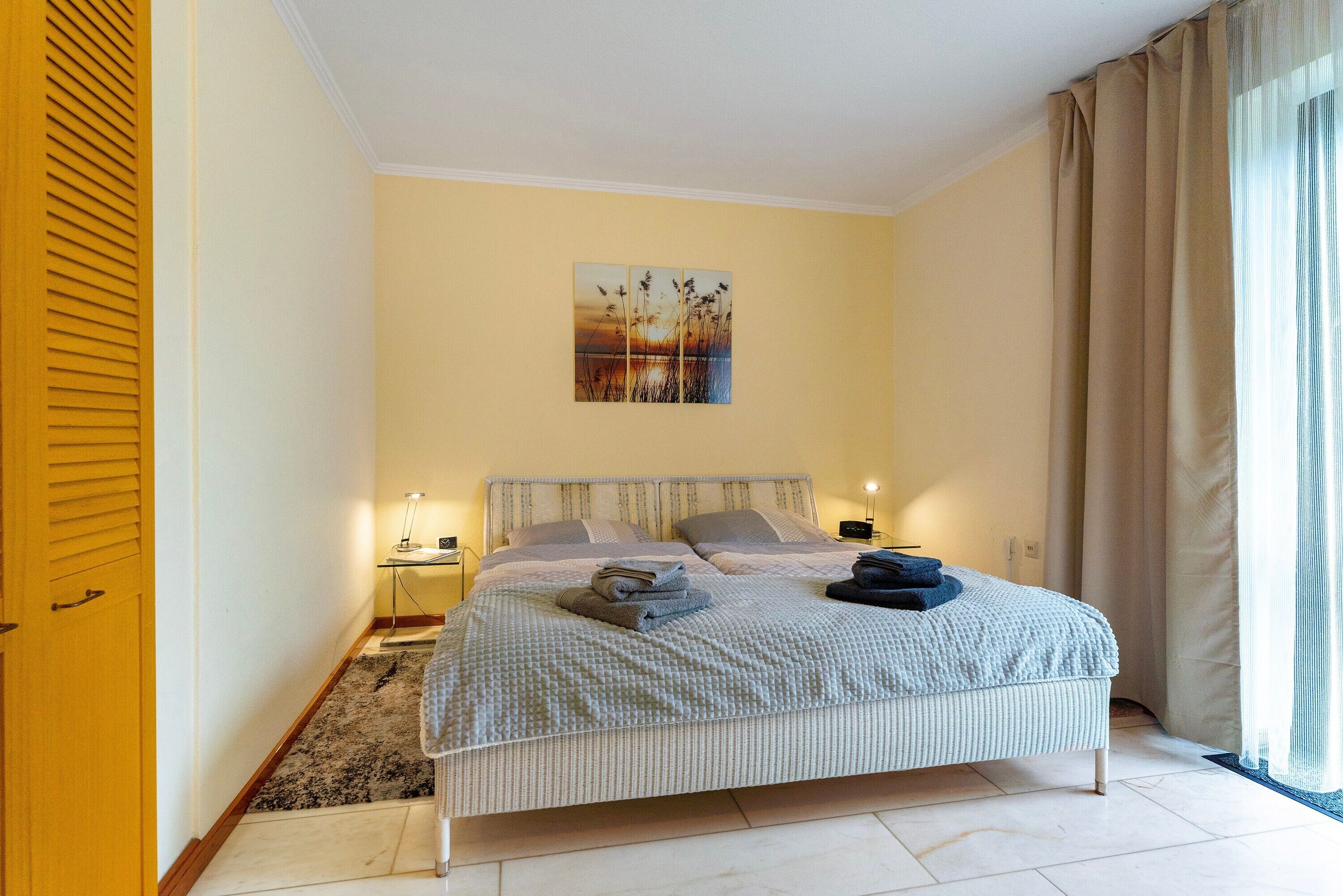 A bedroom with a bed, bedside lamps, a nightstand, a painting on the wall, and a window with curtains.