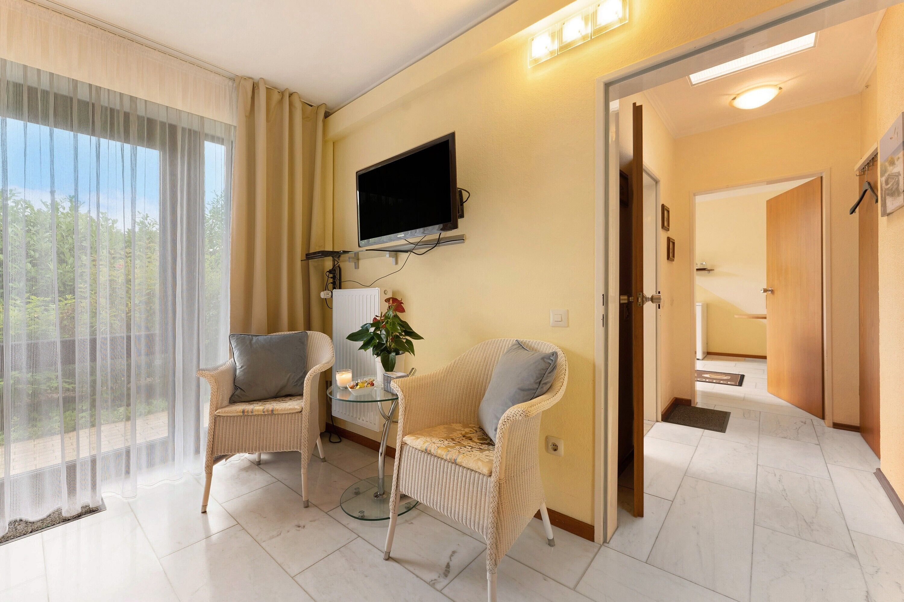 Ein Zimmer mit an der Wand montiertem Fernseher, zwei Korbstühle, einem kleinen Tisch mit Pflanze und Blick durch ein großes Fenster.