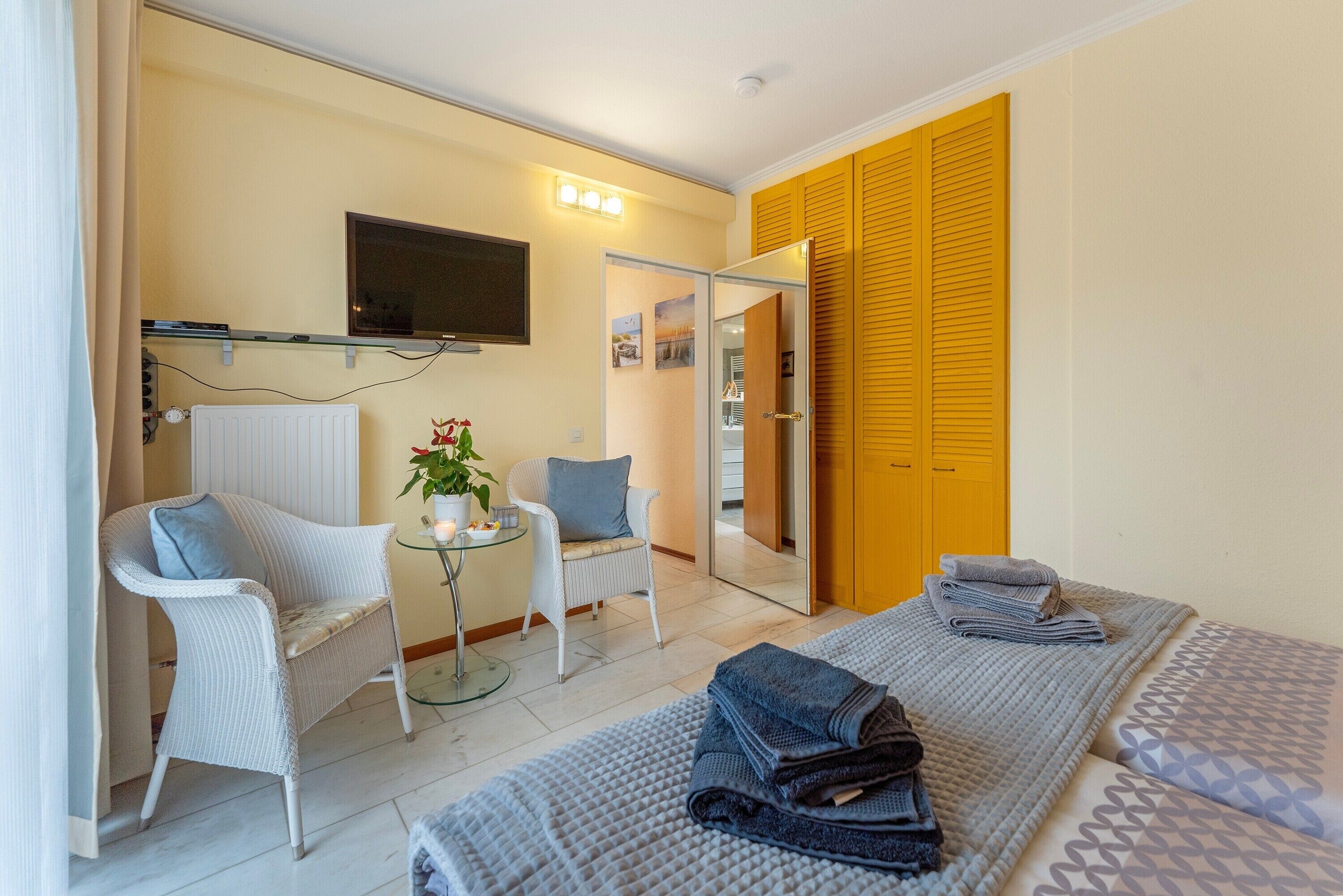 Ein Hotelzimmer mit einem Bett, zwei Stühlen, einem kleinen Tisch mit einer Pflanze, einem an der Wand befestigten Fernseher und einem Schrank mit Schiebetüren.