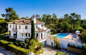 A large white villa with a swimming pool, surrounded by lush greenery and trees.