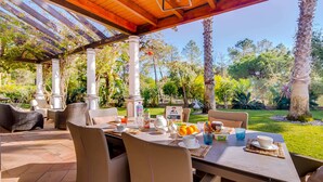 A covered patio with a table set for breakfast, surrounded by chairs, with a view of a garden and trees. - 5 Star Villa Hibiscus - Quinta Verde Retreat (Algarve)