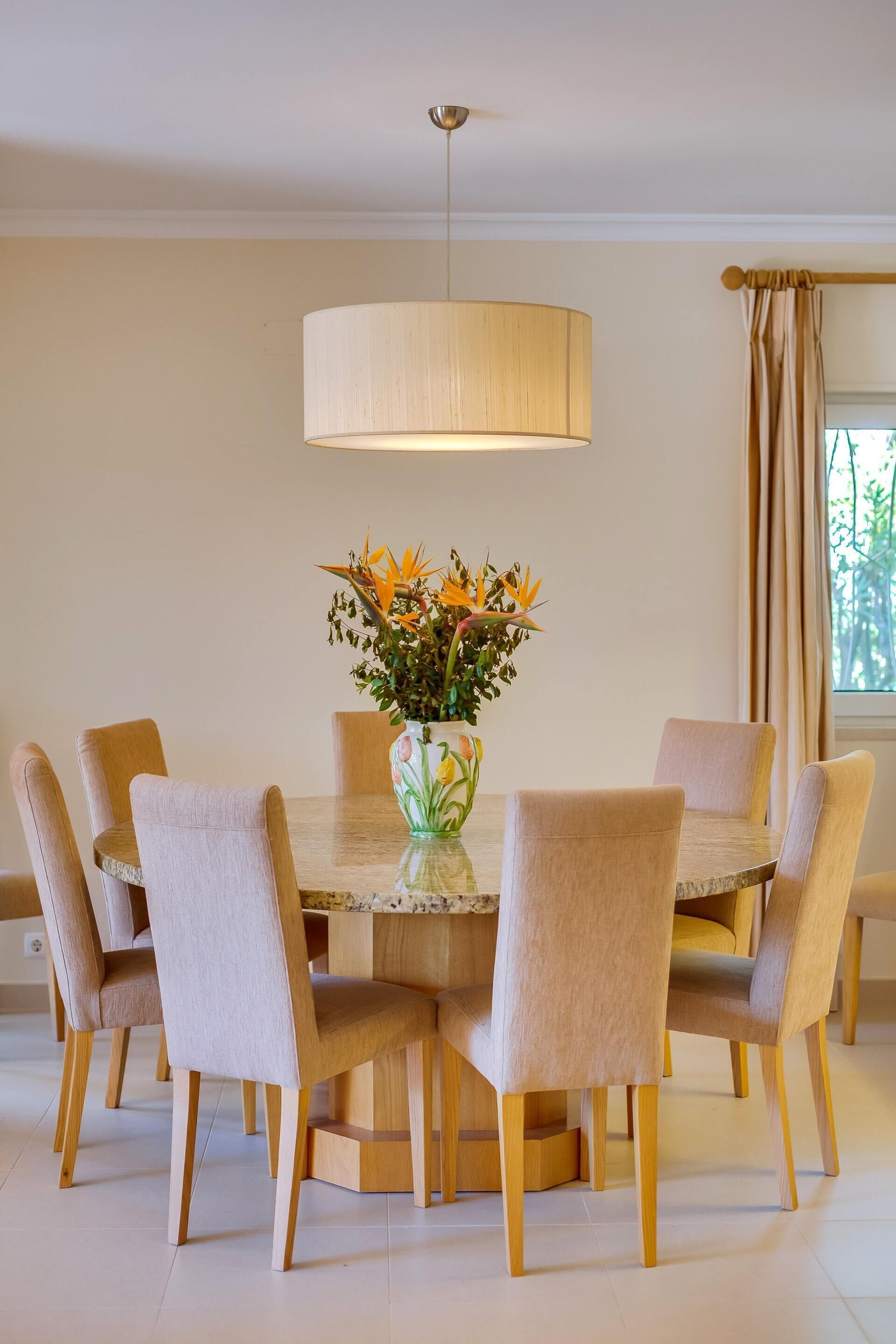 Et spiseområde med et rundt bord, seks stole, en vase med blomster og et hængende lysarmatur.