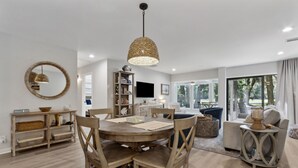 Un salon moderne avec une table de salle à manger ronde en bois, des chaises assorties, un grand luminaire suspendu en fibres tressées, une bibliothèque, un téléviseur à écran plat et un canapé avec une table d’appoint.