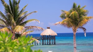 A beach with a thatched-roof structure on stilts over the water, palm trees, and clear blue ocean. - Vidanta Mayan Palace 2 BR 2 BA Suite With Kitchen Sleeps 8 - Cancun Riviera Maya (Carmen Carretera Federal)