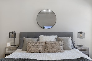 A neatly made bed with a grey headboard, two bedside tables with lamps, and a round mirror on the wall.