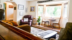 Un salon comprenant un canapé, un fauteuil en cuir, une table à manger avec des chaises, un ventilateur de plafond et une grande fenêtre donnant sur des arbres.