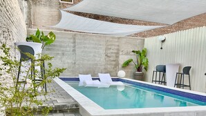Ein rechteckiger Swimmingpool mit weißer Umrandung, umgeben von einer Steinterrasse, einem weißen Baldachin und einer Ziegelwand.