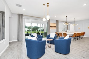 Een moderne woonkamer met blauwe banken, een eetgedeelte met een grote tafel en een uitzicht op een groen landschap door grote ramen.