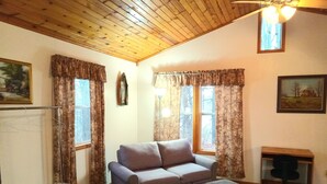 Una sala de estar acogedora con un sofá, un reposapiés y un escritorio con una silla. Hay dos ventanas con cortinas, un ventilador de techo y un cuadro en la pared.