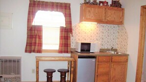 Cocina pequeña con microondas, pileta y armarios de madera. Hay una ventana con cortinas y una mesa con taburetes.