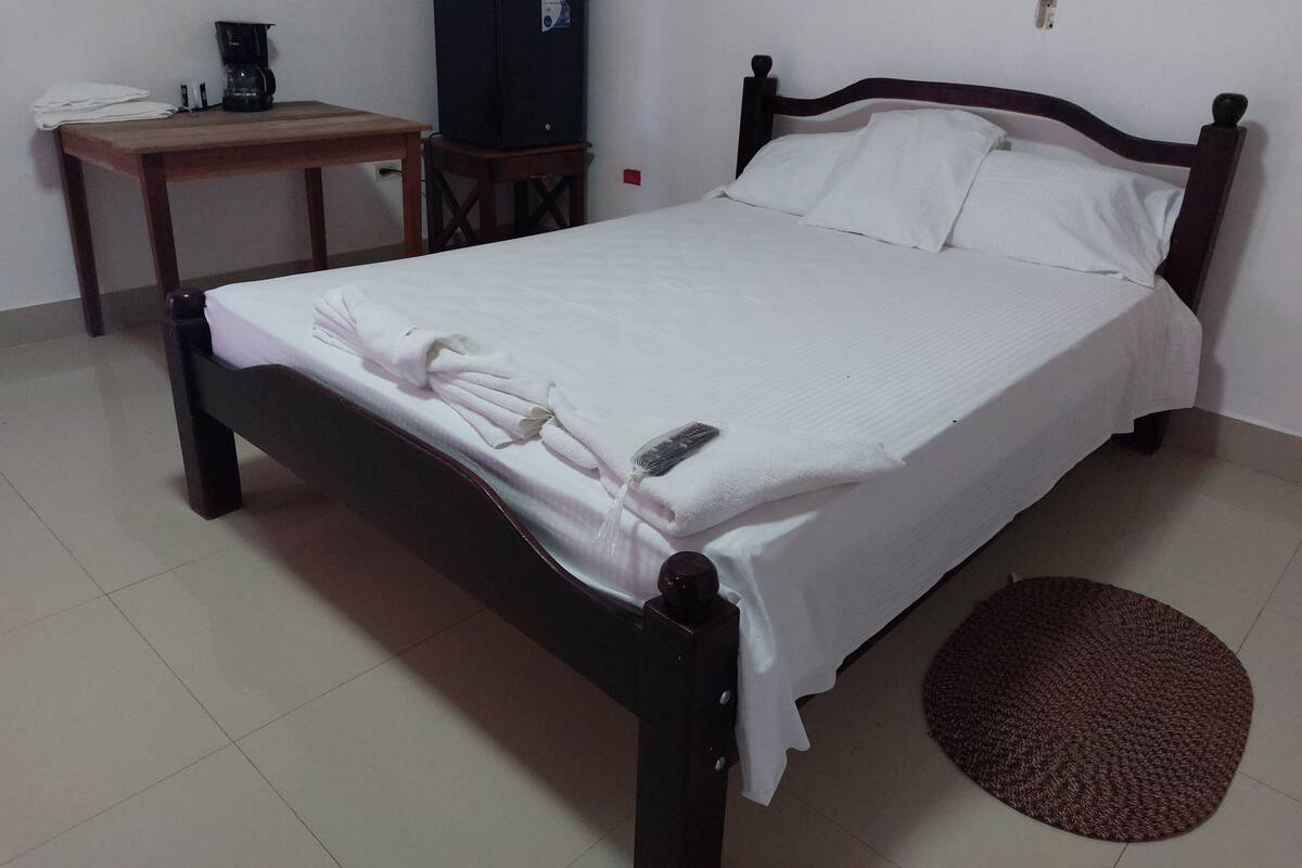 A single bed with white linens, a remote control, and a folded towel on the bed. There is a small wooden table with a coffee maker and a black refrigerator in the background.