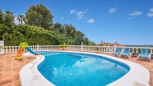 A swimming pool with a slide, surrounded by a white fence and lounge chairs, with trees and a view in the background.