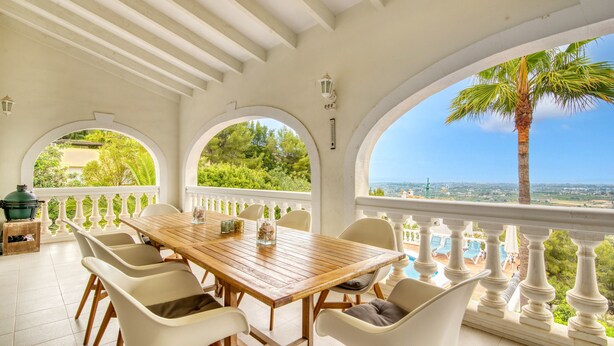 A covered outdoor dining area with a wooden table and white chairs, large arched windows, and a view of a pool and greenery.