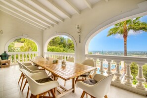 Un espacio de comedor al aire libre cubierto, con una mesa de madera y sillas blancas, amplias ventanas en arco y vistas a una piscina y follaje.