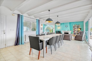 Een eetgedeelte met een lange tafel en stoelen, een keuken op de achtergrond en een boekenkast met boeken.