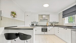 A modern kitchen with white cabinets, a central island with two black bar stools, a built-in oven, and a window with a view of greenery.