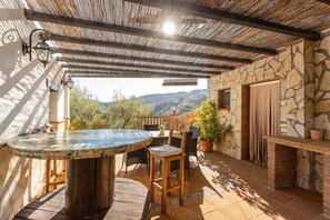 A covered patio with a round table and chairs, stone walls, and a view of the hills. - Holiday Home "Fuente Luz Cottage" with Mountain View, Pool, Wi-Fi, Terrace & Garden (Frigiliana)