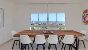 A dining area with a wooden table and white chairs, large windows with a view of the sea and buildings, and a painting on the wall. - Casa La Perla: Villa with Sea View, Pool, Balcony, A/C & Wi-Fi (Benitachell)