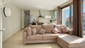 A modern living room with a sectional sofa, a kitchen in the background, and large windows with curtains.