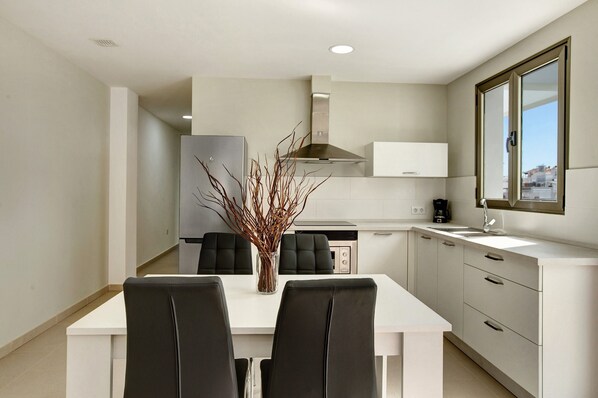 A modern kitchen with white cabinets, a stainless steel range hood, and a dining area with a white table and black chairs. - Apartment Atico Albamar with Mountain View, Wi-Fi, Balcony & Terrace (Puerto de Mogán)