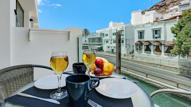A table with a glass of wine, a plate of fruit, and a mug on a balcony overlooking a street with buildings. - Apartment Albamar 1D with Mountain View, Wi-Fi, A/C & Balcony (Puerto de Mogán)