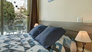 A hotel room with a bed, a nightstand, a lamp, a window with a view of buildings, and a table with chairs outside. - Apartment Albamar 1D with Mountain View, Wi-Fi, A/C & Balcony (Puerto de Mogán)