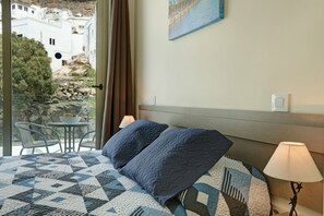 A hotel room with a bed, a nightstand, a lamp, a window with a view of buildings, and a table with chairs outside.