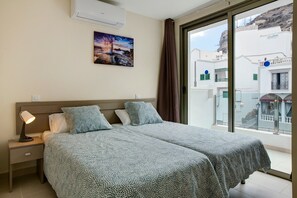 Een hotelkamer met een bed, nachtkastje, lamp, schilderij en uitzicht op gebouwen door een raam.