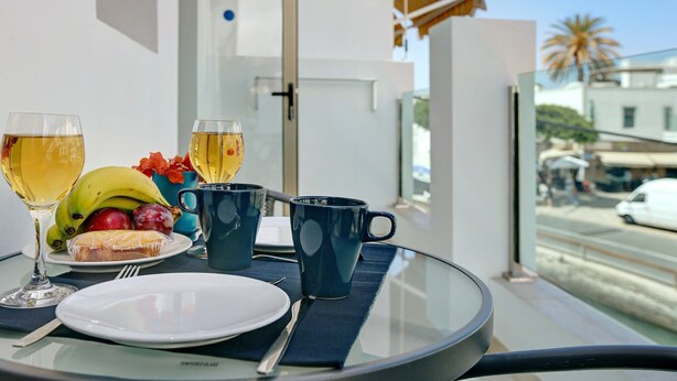 A table with a plate, two glasses of wine, a banana, grapes, and a mug on a balcony overlooking a street. -  Apartment Albamar 1A with Mountain View, Wi-Fi, A/C, Balcony & Terrace (Puerto de Mogán)
