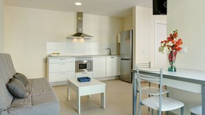 A modern kitchen with white cabinets, a stainless steel range hood, a built-in oven, and a refrigerator. There is a dining table with chairs and a small white table with a blue basket. A grey sofa with cushions is visible in the foreground. -  Apartment Albamar 1A with Mountain View, Wi-Fi, A/C, Balcony & Terrace (Puerto de Mogán)