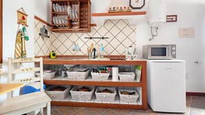 Cocina equipada con microondas, lavamanos y diversos utensilios de cocina. Se observan cestas de mimbre en estantes y un armario de madera con vajilla.