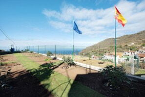 Un espacio al aire libre cercado con vistas al mar, una bandera y un paisaje montañoso al fondo.