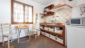 Una cocina compacta con microondas, repisas con vajilla, una silla, una mesa y un ventanal con cortinas.