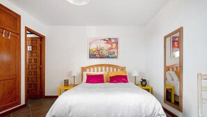 Un dormitorio con cama de madera, ropa de cama blanca, un cuadro colorido, un espejo y un armario de madera.