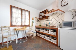 A compact kitchen with a microwave, shelves with dishes, a chair, a table, and a window with curtains.