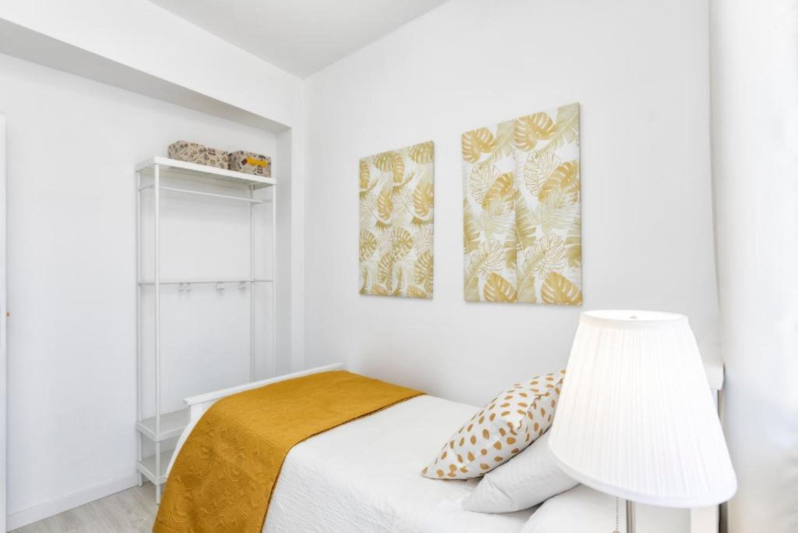 Een slaapkamer met een bed, een witte lamp, een plank met decoratieve objecten en twee kunstwerken aan de muur.