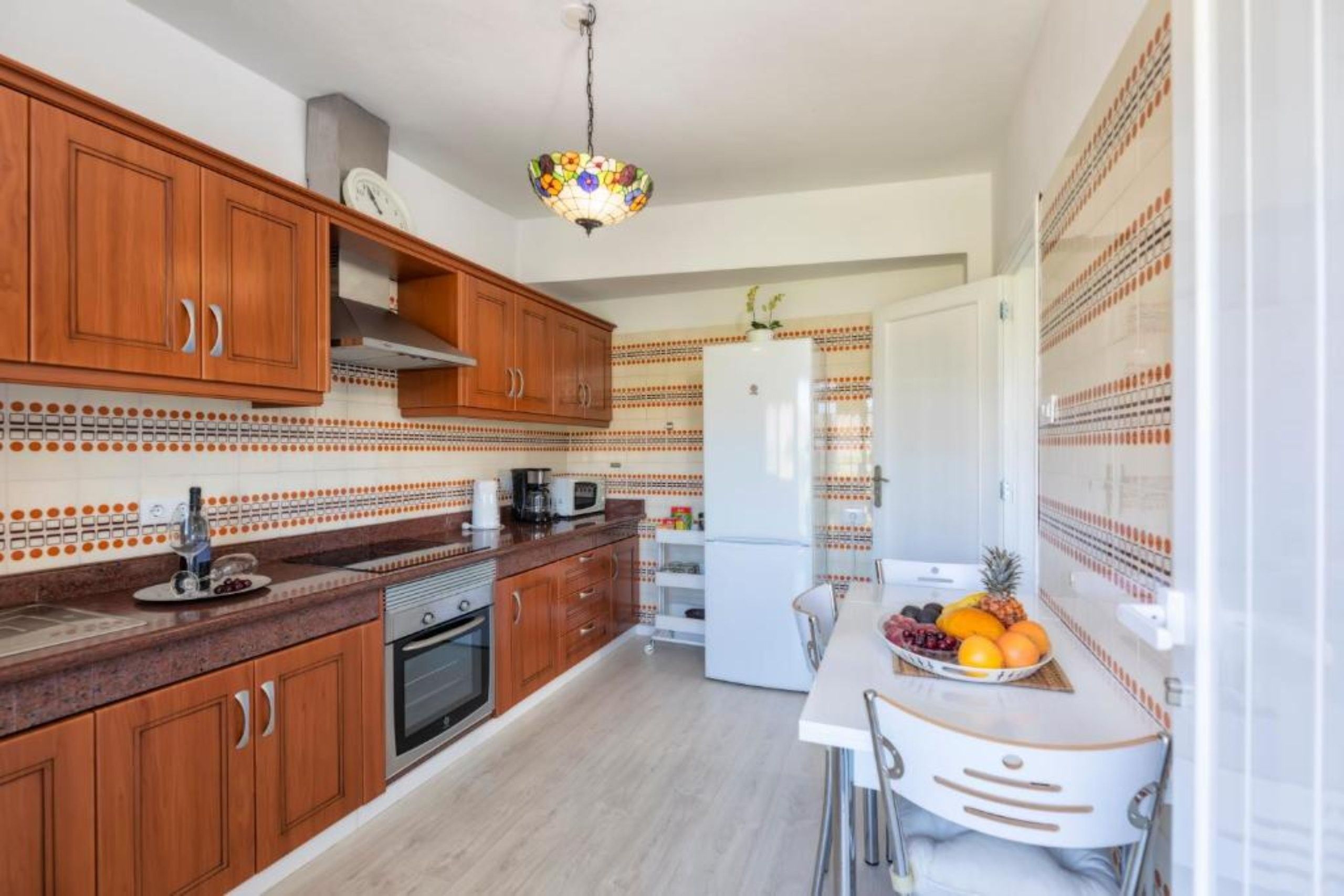 A kitchen with wooden cabinets, a white refrigerator, a dining table with chairs, and a fruit bowl.