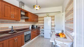 A kitchen with wooden cabinets, a white refrigerator, a dining table with chairs, and a fruit bowl.