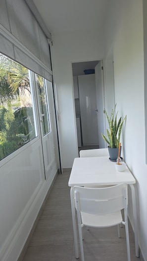 Un pasillo estrecho con una mesa y sillas blancas, una planta en maceta y vistas de vegetación al exterior.