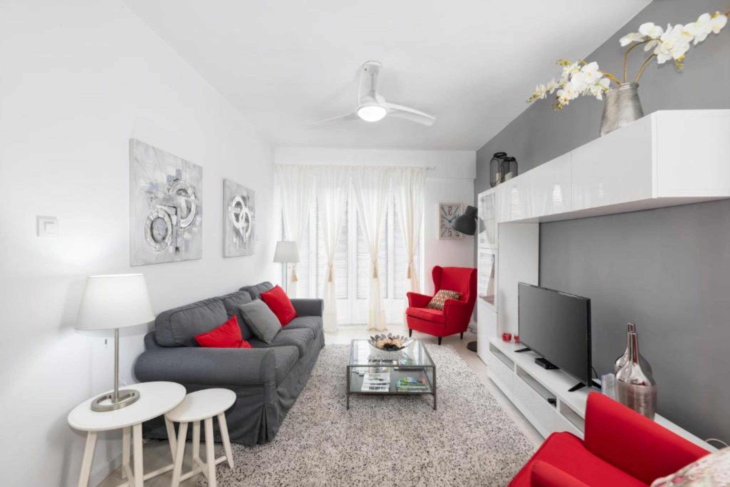 Ett modernt vardagsrum med en grå soffa, en röd fåtölj och en platt-TV. Det finns en takfläkt, ett soffbord och ett litet sidobord med en lampa.