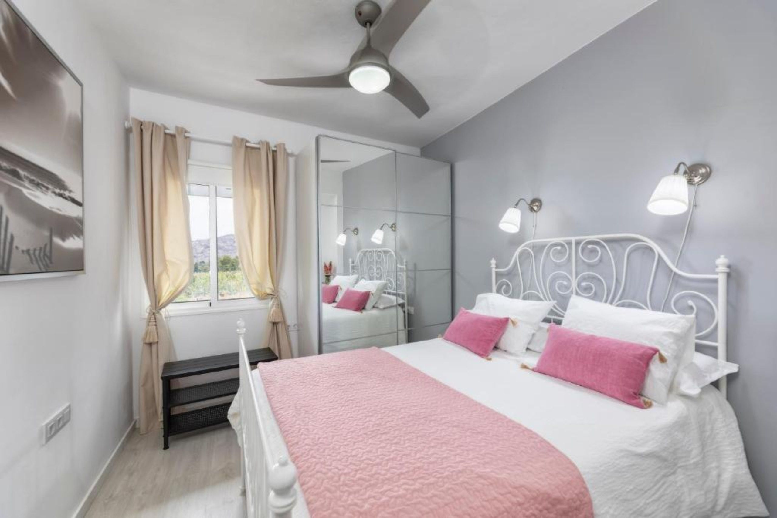 Une chambre à coucher comprenant un lit, un ventilateur de plafond, une fenêtre avec des rideaux, un miroir et des lampes fixées au mur.