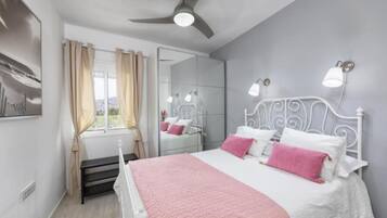 Une chambre à coucher comprenant un lit, un ventilateur de plafond, une fenêtre avec des rideaux, un miroir et des lampes fixées au mur.