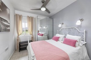 A bedroom with a bed, a ceiling fan, a window with curtains, a mirror, and wall-mounted lights.