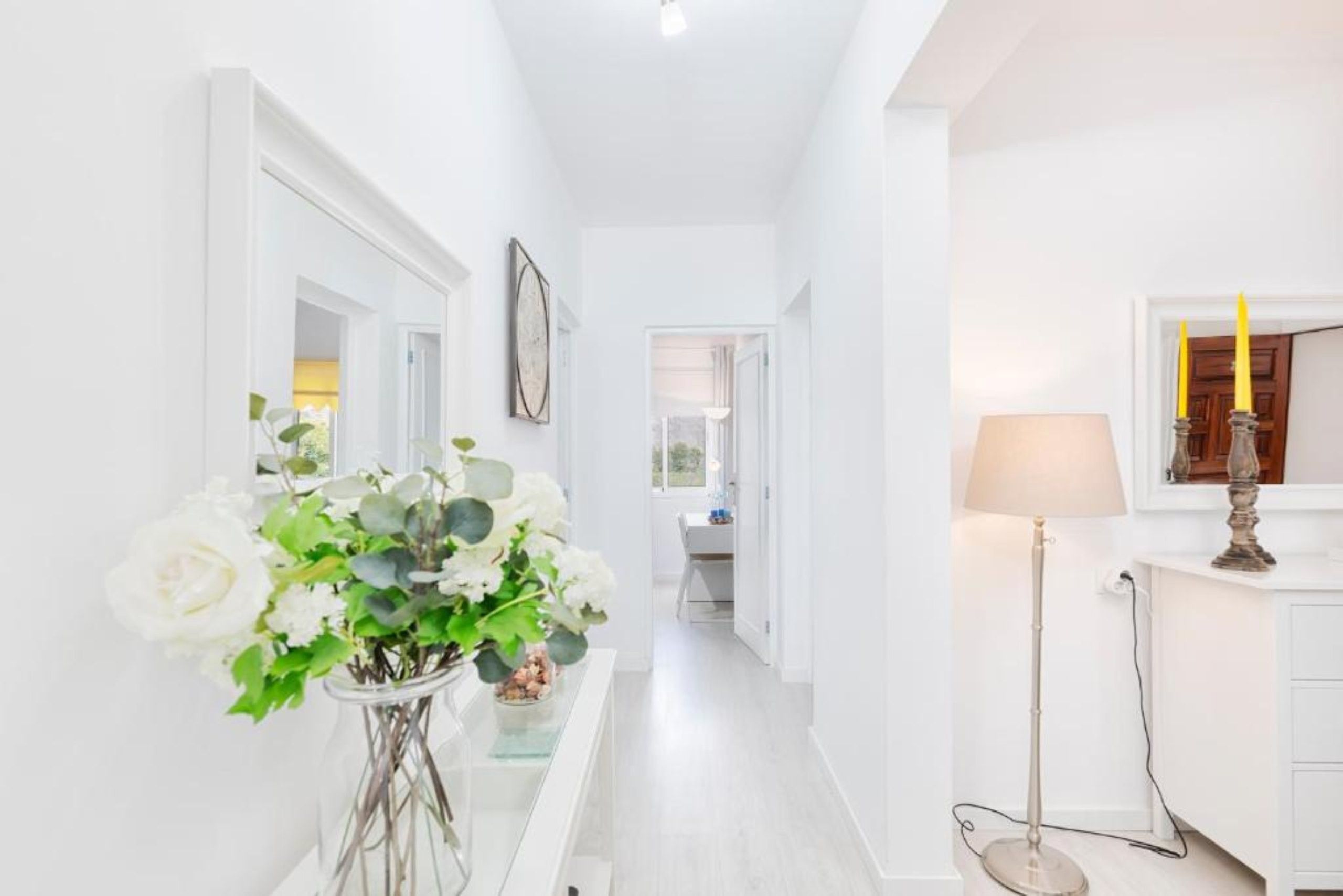 Un pasillo con un jarrón de flores blancas, una lámpara y un espejo en la pared.