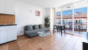 Un salon avec un canapé, une table basse en verre, un coin repas avec des chaises, une cuisine avec un mur de crédence en briques et un balcon offrant une vue sur les immebles.