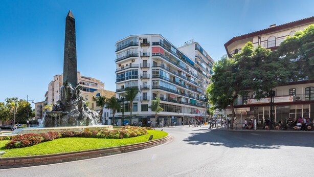 Une place publique agrémentée d’une fontaine, d’un monument imposant et d’immeubles à plusieurs étages dotés de balcons.