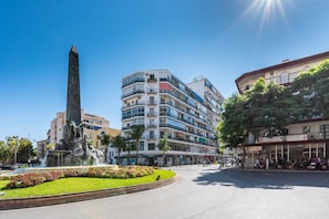 A public square with a fountain, a tall monument, and multi-story buildings with balconies.