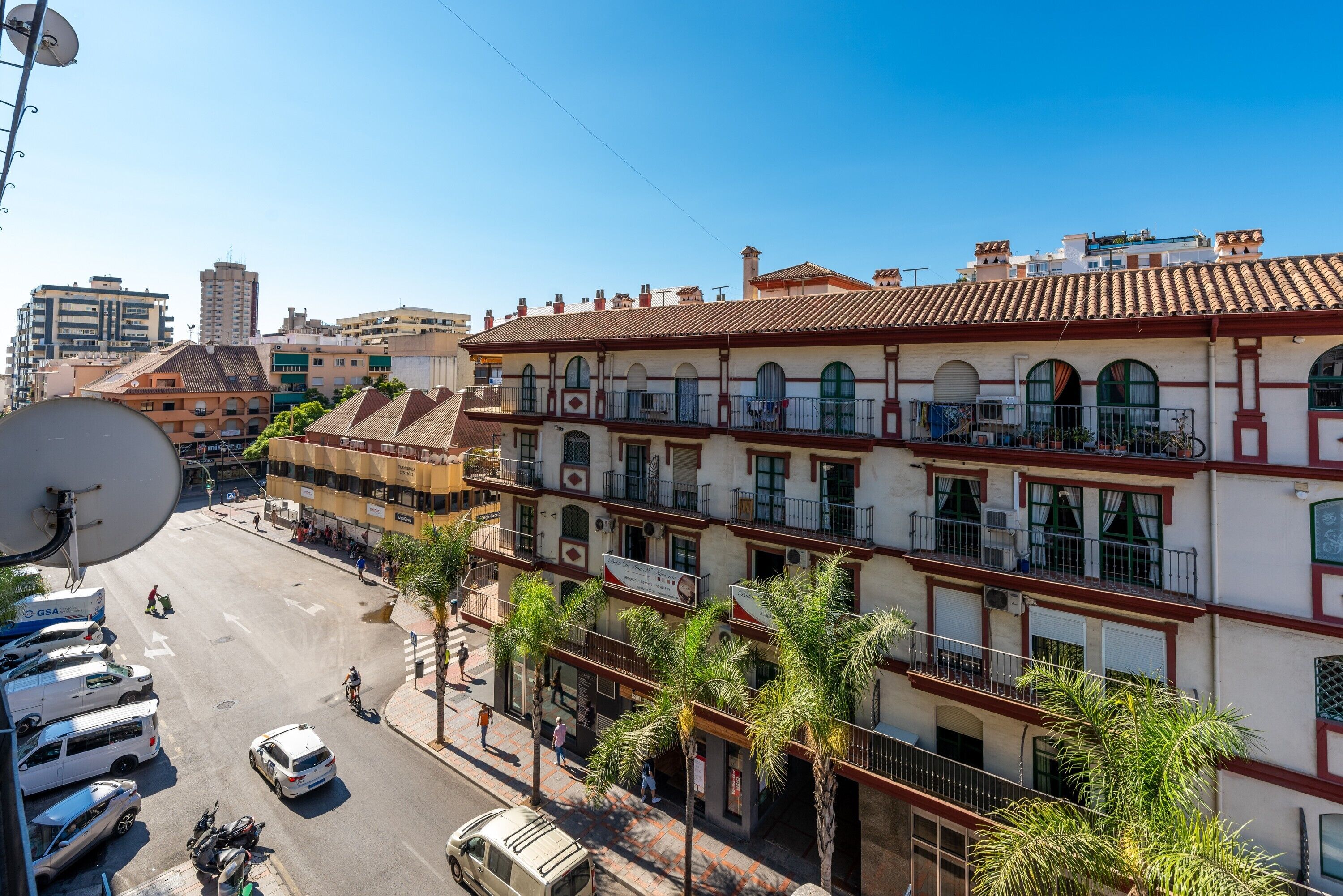 A multi-story building with balconies, a street with vehicles, and palm trees.