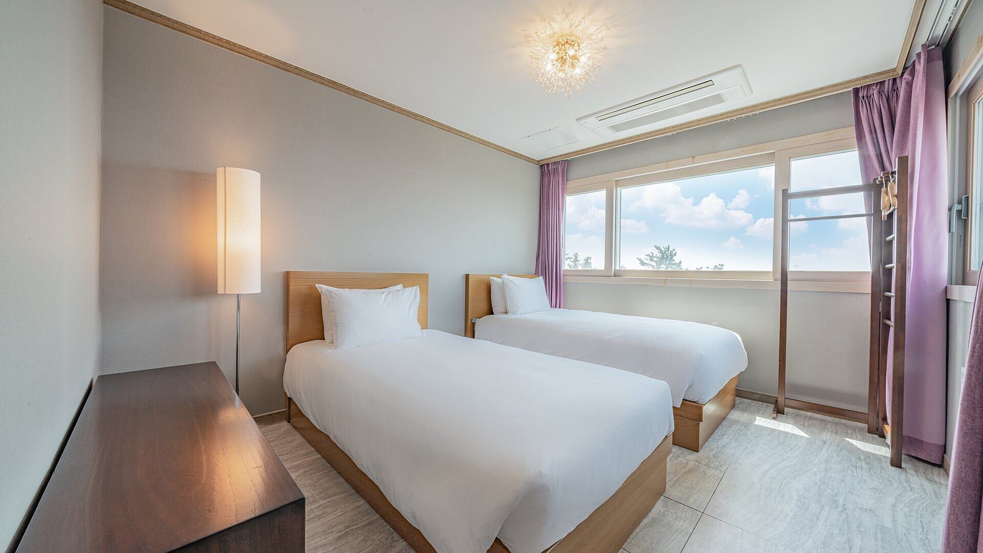 Habitación de hotel con dos camas, una mesita de noche, una lámpara, una ventana con cortinas e iluminación en el techo.