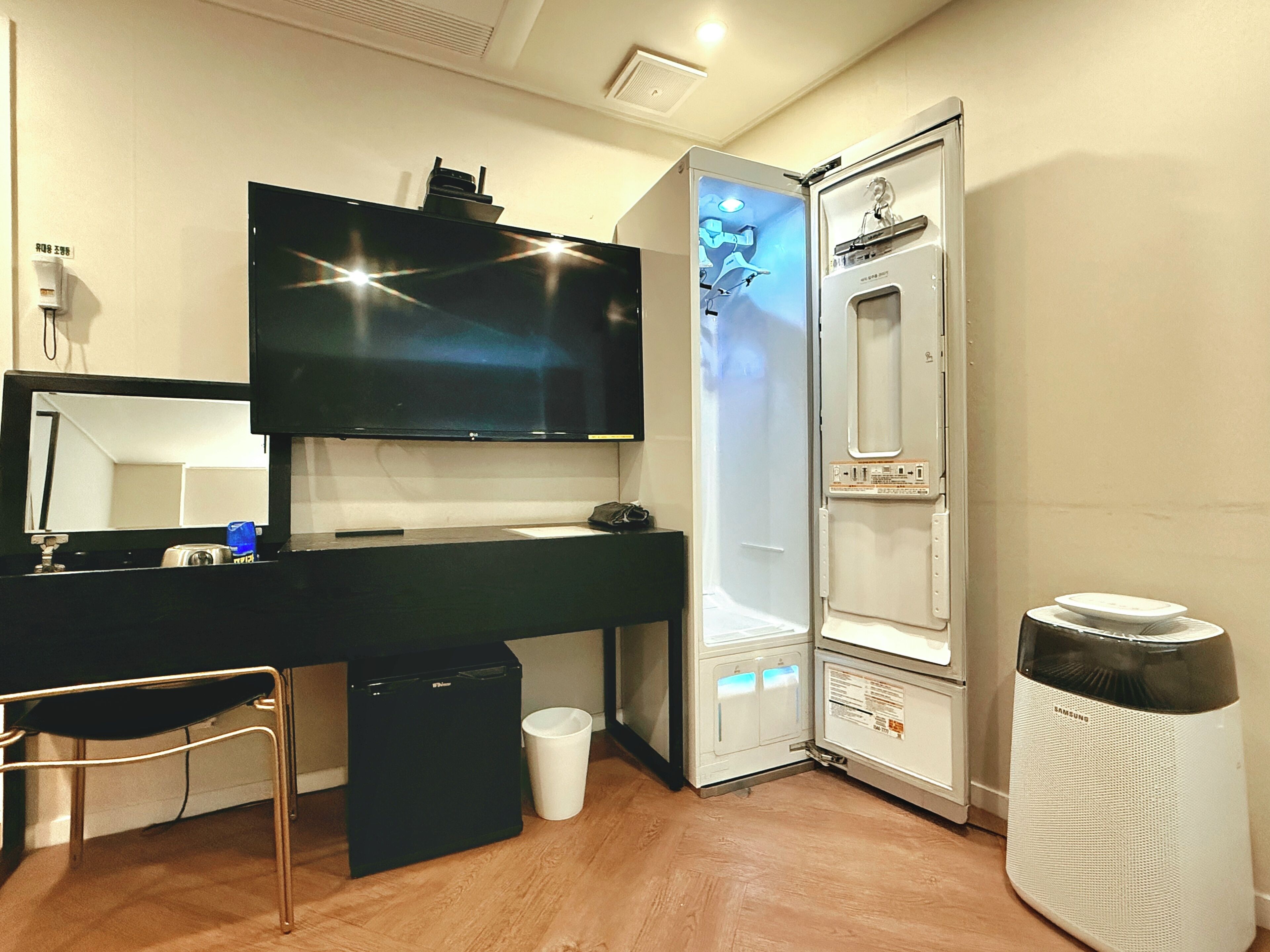 Una habitación con un escritorio negro, un televisor montado en la pared, un cubo de basura blanco y un purificador de aire blanco.